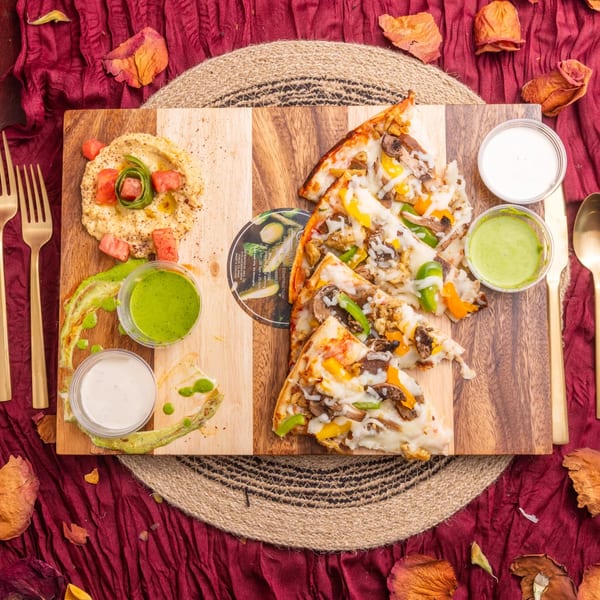 Hummus, vegetable pizza slices, and dipping sauces on a wooden board with gold cutlery.