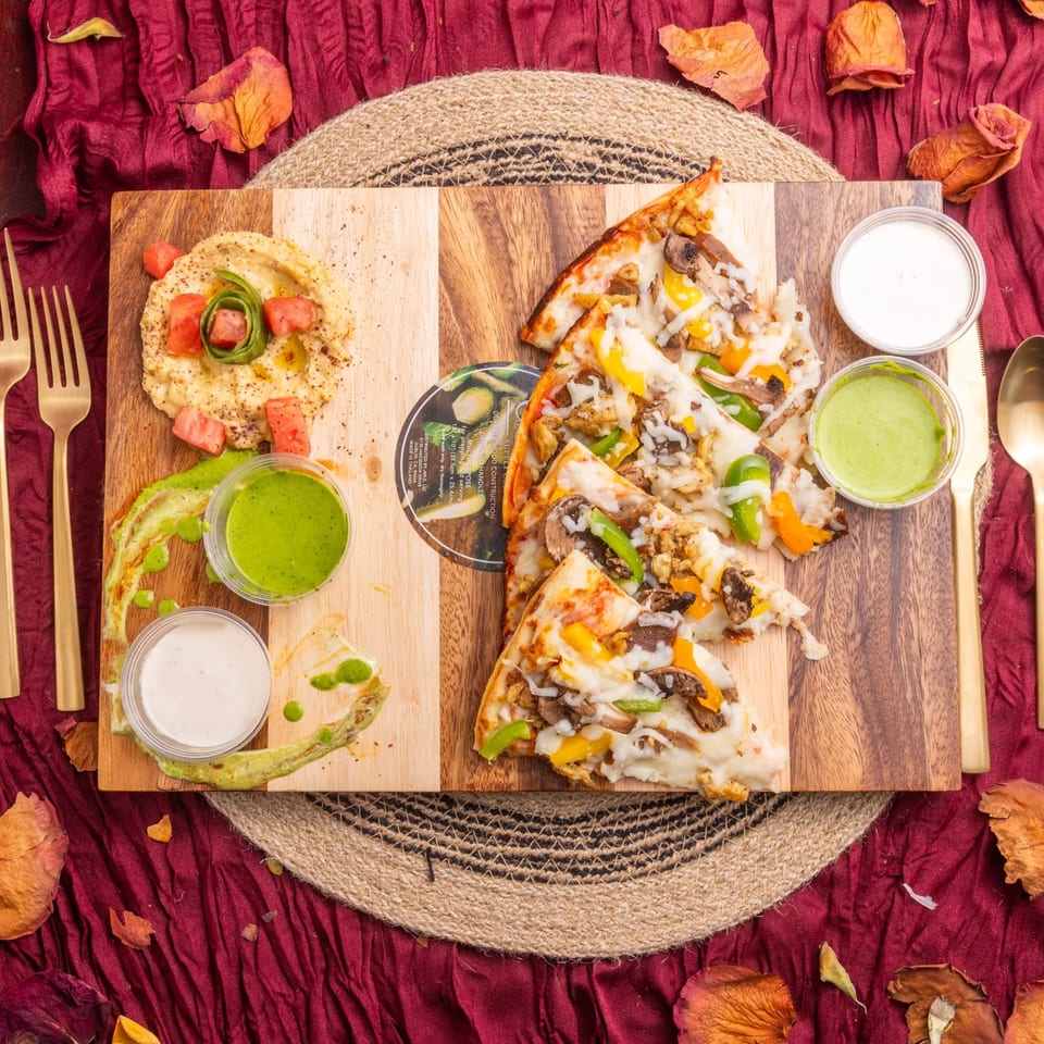 Hummus, vegetable pizza slices, and dipping sauces on a wooden board with gold cutlery.