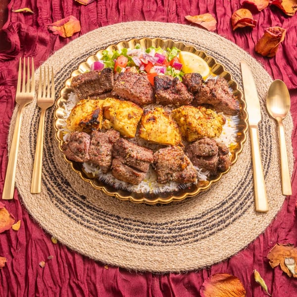Golden platter of beef and chicken kebabs over rice, with salad and lemon, on a woven mat.