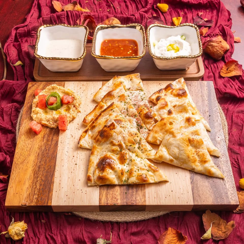 Hummus with pita bread and three dipping sauces on a wooden board with a red cloth.